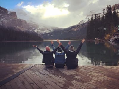 British Columbia, here we come! Drei Studentinnen sitzen am Rand eines Sees mit spektakulärer Wald- und Gebirgskulisse im Hintergrund