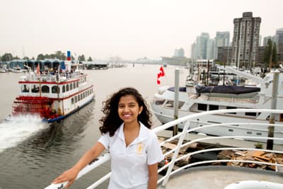 Entdecken und selbst mitanpacken! Tourism Management Studentin an Deck eines Schiffes. Im Hintergrund fährt ein Raddampfer vorbei und man sieht die Hochhäuser der Innenstadt Vancouvers.