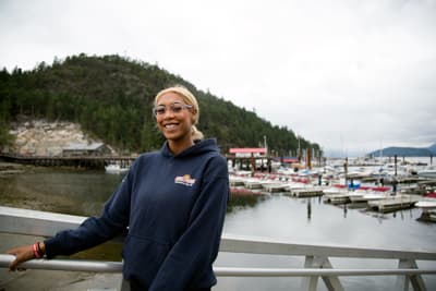 British Columbia entdecken Eine Tourism Management Studentin vor dem Hintergrund eines bewaldeten Hügels am Wasser und eines Hafens.