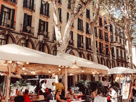 Ein Semester lang Dolce Vita! Sommerliche Caféterasse mit Lichterkette im Sonnenschirm vor einer schönen Altbaufassade und unter Platanen in Barcelona.