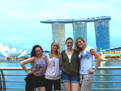 Genieße den Lifestyle Singapurs Internationale Studentinnen posieren an der Uferpromenade vor dem Panorama des leuchtenden Marina Bay Sands Hotels
