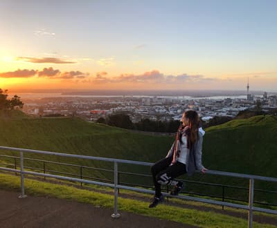 Action und schlafende Vulkane in Auckland Studentin posiert auf einem Geländer am grünen Mount Eden. Im Hintergrund sieht man die Stadt Auckland aus der Ferne mit ihrem charakteristischen Sky Tower.