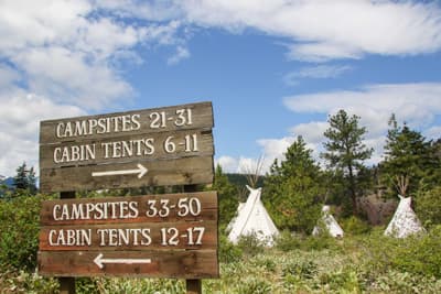 Probieren geht über Studieren! Tipi-Zelte vor einem Wald und ein Wegweiser, der die Richtung zu bestimmten Lagerplätzen des Campingplatzes weist.