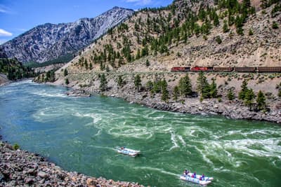 Action oder lieber entspannt? Reißender türkisfarbener Fluss, auf dem zwei Rafting-Boote fahren vor einer Berglandschaft mit Zug