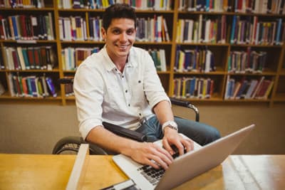 Mit Behinderung ist eine gute Organisation besonders wichtig. Ein Rollstuhlfahrer sitzt in einer Bibliothek am LAptop.